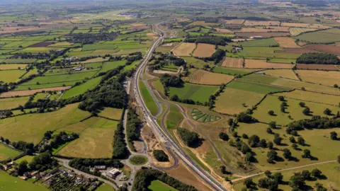 National Highways An aerial shot of the A303 between Sparkford and Ilchester.  There are green fields and trees either side of the road.