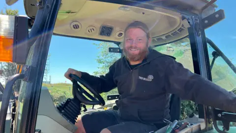 A picture of James sat inside a vehicle. He has a ginger beard and is wearing a black jumper.
