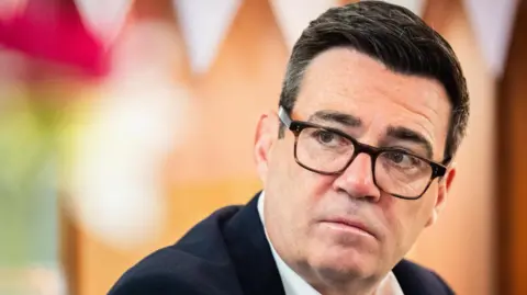 PA Andy Burnham, wearing a suit and glasses, is looking off camera with a serious expression. 