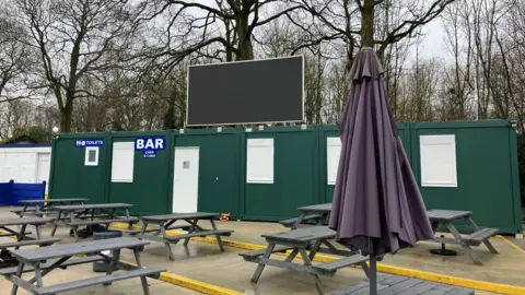 A set of picnic tables and a large umbrella on a concrete deck. Behind it, green cabins that have signs for a bar and toilets, and above those a large screen. In the background, some trees without their leaves