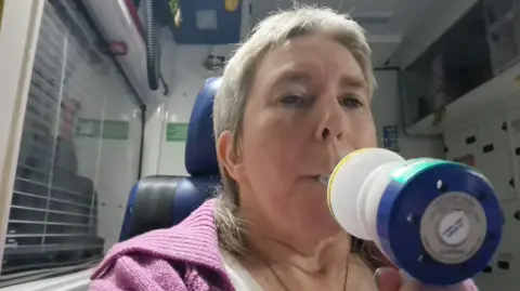 Helen Parry Parry takes a selfie in an ambulance while taking on gas and air for the pain. She has short grey hair and is wearing a pink cardigan. 