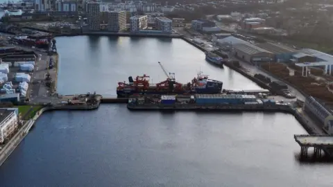 A large vessel docked at a quayside at Chatham Docks. The water in the harbour, which is surrounded by commercial buildings and blocks of flats, appears still.