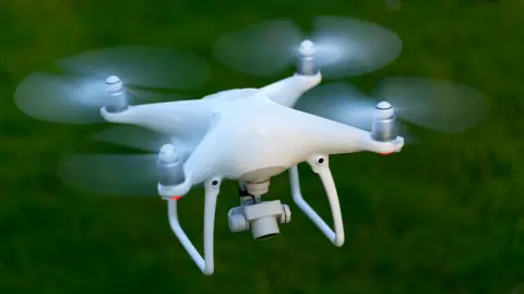 A white drone with four propellors and a camera looking down underneath it