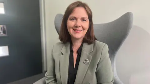 Woman with short brown hair in green linen jacket with black blouse and silver broach sits in grey armchair 