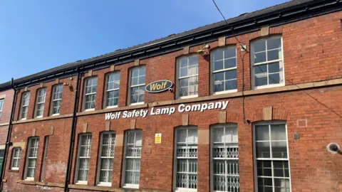 BBC/Jack Hadaway-Weller A large red brick building with two rows of windows. The words "Wolf Safety Lamp Company" stretch in white letters across the bottom row of windows. A smaller, round logo with the word "Wolf" is above.