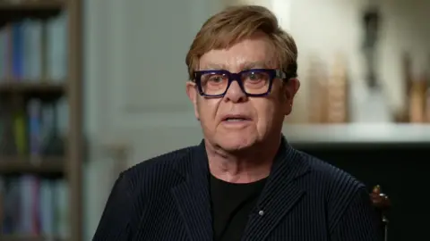 Sir Elton John speaking to Laura Kuenssberg, who is behind the camera. He has ginger hair and blue eyes. He is wearing a black t-shirt, black blazer, and thick, square, black-rimmed glasses.