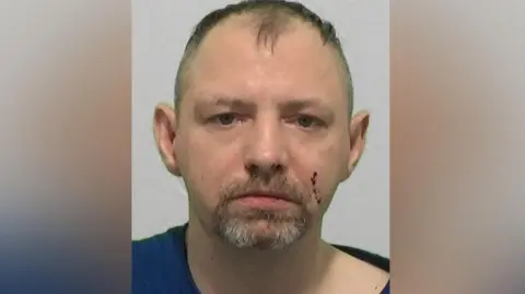 Northumbria Police Mugshot of Ian Hudson. He has a small brown and grey goatee and thinning fair hair, with cuts on his cheek and nose.
