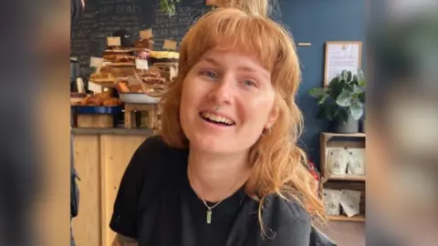 Ruby Love, wearing a black t shirt and necklace, smiling at the camera. She also has a septum piercing and wears her long strawberry blonde hair down with a fringe. Behind her are what looks like cake and displays that show a cafe. 