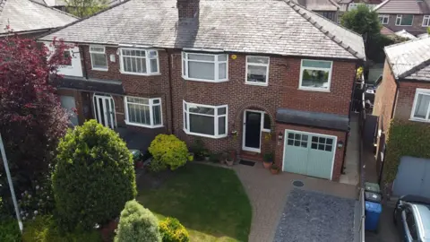 A brown brick semi-detached house with a light green garage door and a front garden with a lawn, trees and bushes.