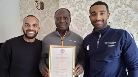 Nelson is stood in between two men who both have dark hair and beards. One is wearing a black jumper and the other is wearing a navy blue sports top with Liverpool John Moores University written on it in white writing.