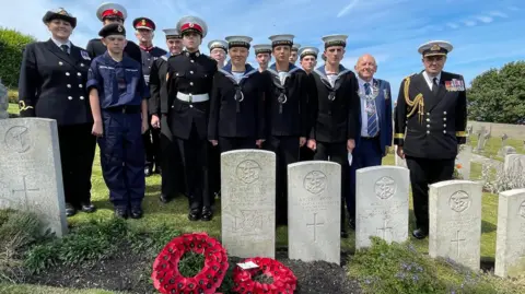 Portland Port Dignitaries stand at the grave of Leading Seaman Jack Mantle on the 85th anniversary of his death during a German dive bomber attack on Portland Harbour on 4 July 1940. They included HM Lord-Lieutenant of Dorset Michael Dooley, the Mayor of Portland Town Council Charlie Flack, Portland Sea Cadets and Yeovil Sea Cadets from TS Mantle VC, named in honour of Jack Mantle. There are poppy wreaths laid at the grave. It is a clear day.