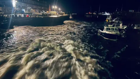 Bridport Harbour / Dorset Council White water rushing through the sluice gates at West Bay at night. Small boats can be seen at the edge of the harbour and the quayside is lit by floodlights.