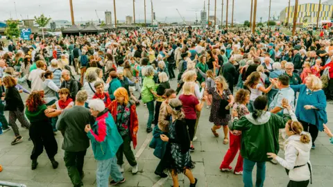 TradFest A very large crowd of people are dancing. Some trees, poles and buioldings are in the background. 