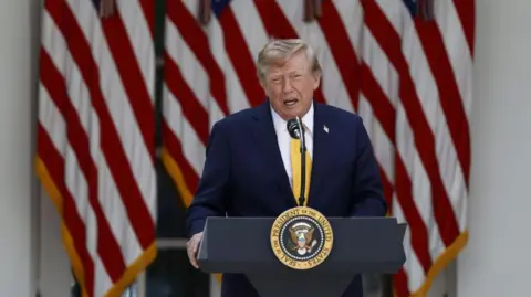 US President Donald Trump delivers remarks outside the White House. He is speaking at a podium with the US presidential seal at the front. He is wearing a dark navy blue suit and a yellow tie. Behind him are several US flags. 