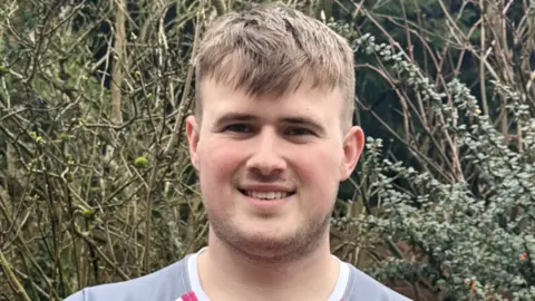 Samuel is standing outside by a hedge in a pink and grey sports top. He is smiling at the camera and has short brown hair a short beard.