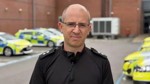 Inspector Joe Senior is pictured standing outside a police station. He is wearing a black police uniform and a pair of glasses. He has grey receding hair. There are several police cars parked in the station car park in the background.