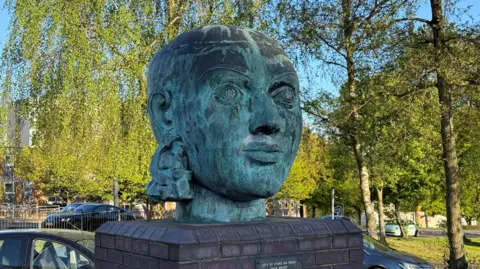 A green-coloured sculpture of a head on a brick plinth is situated at the edge of a car park. There are trees and a road in the background behind it.