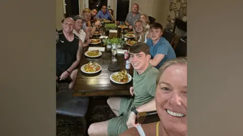 A group photograph of family and friends sat around a table having a meal.