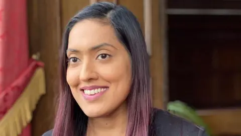 Sahida Ditta with long dyed red hair and a black top. She is smiling. The background is blurred.