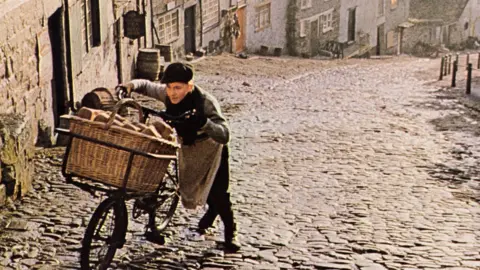History of Advertising Trust A colour still from an advertisement for Hovis. It shows a baker's boy, wearing a cap and long apron, pushing a bike with a large wicker basket full of loaves of bread up a very steep cobbled street. There are traditional looking cottages on the right hand side.