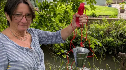 A woman with short brown hair and brown-rimmed glasses, and wearing a light blue and white striped top, is on the left hand side of the image looking towards the camera. On the right ahnd side her left arm is holding up a small stainless steel bucket on a red string, used for collecting water samples. The end of a domestic garden, a row of foliage and river Isbourne are visible in the background.
