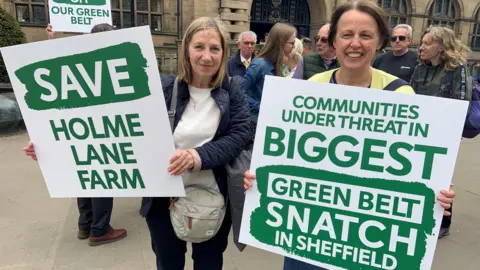 Two women are standing outside Sheffield Town Hall holding green and white placards protesting the decision to build on green belt land