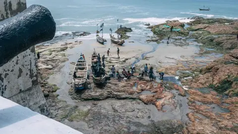 AFP via Getty Images Fishermen tending to boats on the shore of Cape Coast Castle. The picture is taken from the castle. To the left of the shot is a partial cannon
