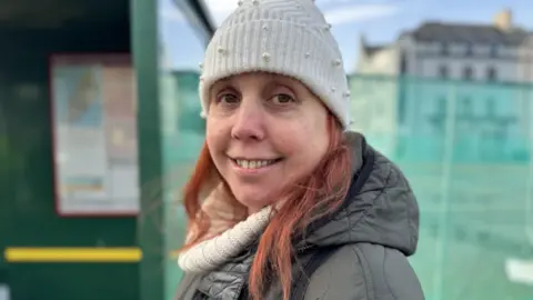 Kim Hidson is smiling wearing a white woolly hat with her red hair at the sides. She is wearing a woolly jumper and waterproof coat and is standing in front of a green bus shelter.