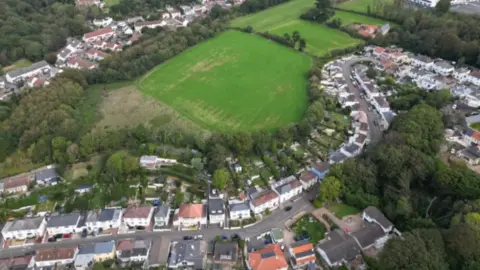 BBC Aerial shot of properties in Jersey. A road runs through a row of houses. A large green field is behind the properties.
