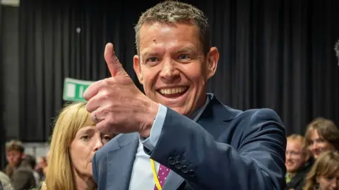 Getty Images Leader of Plaid Cymru Rhun ap Iorwerth gives a thumbs up sign during the count in Llandudno. Dressed in a dark blue suit.