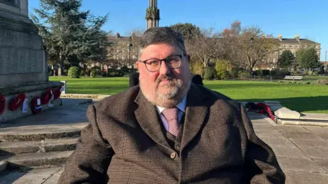 Philip Barton, age 57, wearing a brown suit, in Hamilton Square, with paving and a green lawn seen behind him.