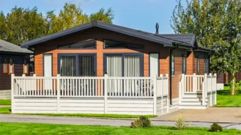 A single-story caravan-lodge with a wooden exterior and a dark grey pitched roof. The facade includes large rectangular windows fitted with vertical blinds. A small porch with wooden steps leads up to the front door, which is partially visible. The house is enclosed by a white picket fence, and the surrounding yard is neatly maintained with green grass. In the background, there are a few trees, suggesting a suburban or semi-rural setting.