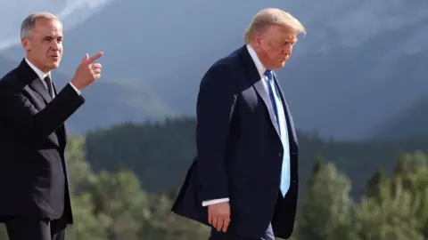 Canada's Prime Minister Mark Carney walks behind US President Donald Trump as they attend a family photo session during the G7 summit in Kananaskis, Alberta. Both wear dark suits, and Carney smiles and gestures while Trump frowns slightly. 