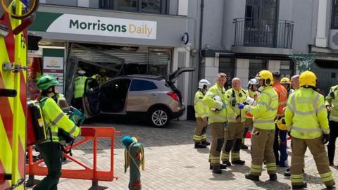 Emergency services in high visibility clothing can be seen gathering around the front of a Morrisons Daily in Romsey where a silver car can be see crashed into the shop front