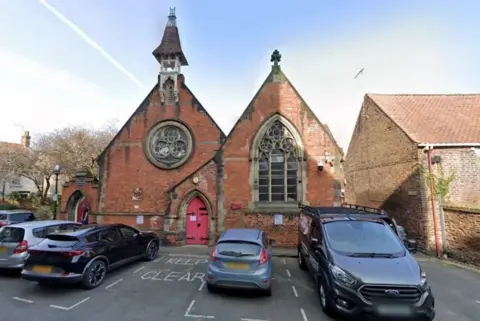 Beverley Minster Parish Hall, with its bell tower. There are cars parked in front of the red brick building.
