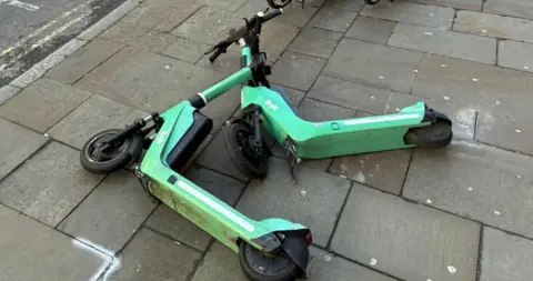 Two green e-scooters discarded on the pavement