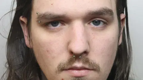 A police mugshot shows a man with long brown hair, a moustache and a goatee style beard, along with piercings in his nose and his right eyebrow.