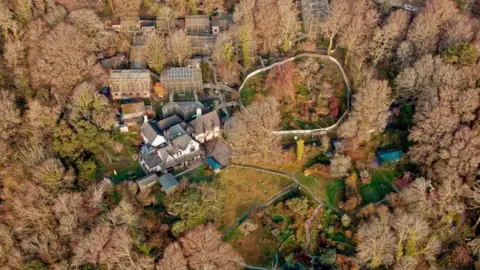 Wild Futures A birds eye view shot of orange and brown trees. In the middle there is a clearing of a huge house and various other cage and brick type buildings.