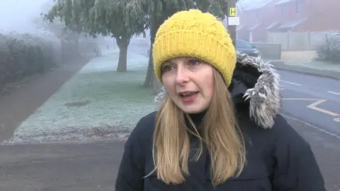 Steve Humphrey/BBC Ros Finney standing next to a road on a frosty day. She wears a yellow knitted hat and black parka coat. Behind her is a pavement and grassy verge with trees, and there are school road markings on the road.