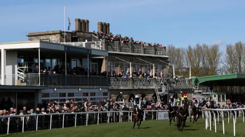 Getty Images A packed grandstand at Kelso races with people also lined up behind the running rail watch as horses stream across the finish line