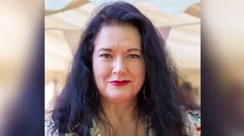 Image shows Ann Millington looking at the camera, she has long dark hair and is wearing a floral top