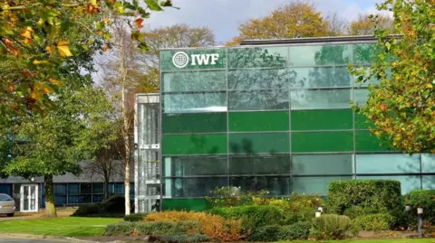 IWF Internet Watch Foundation's offices are a three-storey glass building with bushes and trees around the, and a car parking area to the left