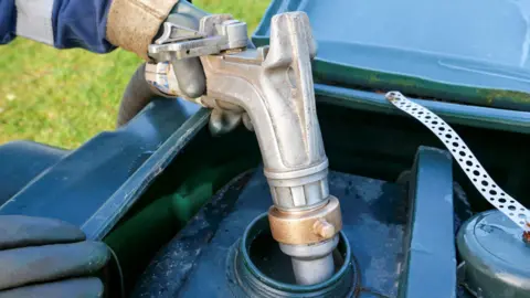 Getty Images A delivery driver places a metal nozzle into the open lid of a heating oil tank to refill it. The driver is wearing protective gloves and overalls.