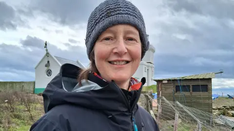 RSPB Sarah Dalrymple wearing a waterproof coat and a woolly hat. She is standing on Coquet Island. A shed and the island's lighthouse stands behind her.