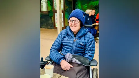 Amy Attwood A man wearing glasses, a navy blue beanie hat and a blue puffer jacket sits smiling in a powered wheelchair at an outdoor table. A cup of tea or coffee sits on the table in front of him. Other people can be seen sitting at tables in the background.