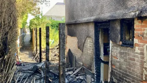 The side of a bricked-house which is blackened with smoke damage. There is charred debris on the ground spilling into an alleyway adjoining the property. Fence panels have clearly burned through.