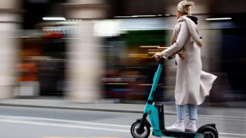 A woman riding on an e-scooter on the road. She is wearing a long cream-coloured coat and scarf, blue jeans and white high-top trainers. She has blonde hair clipped back with a clasp.