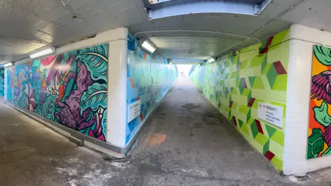 Image shows the Bedminster station underpass from a different angle, with a path leading to platform one. The path stretching up to the platform has one green and one blue wall, with varying shades and patterns. 