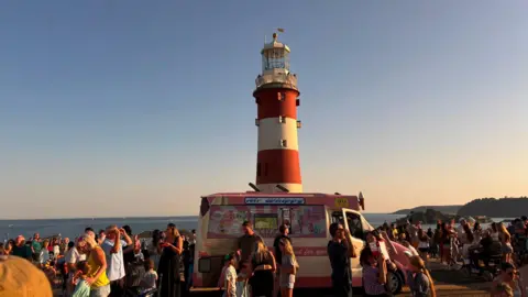 A picture of an ice cream van on Plymouth Hoe. There is a number of people crowded around in front of Smeaton's Tower.