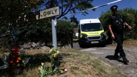 A police officer is seen walking beneath a cordon with a police van in the background and to the left a Drive Slowly sign with flowers underneath it.